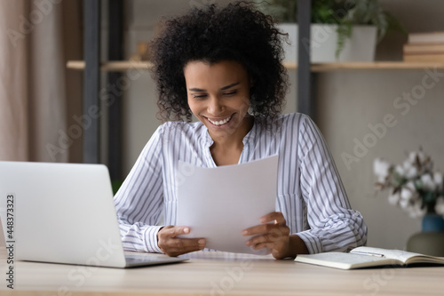 Happy young African American woman sit at desk at home office work online on computer with paperwork documents. Smiling biracial female read good news in paper correspondence. Success concept.
