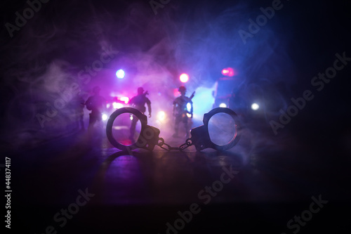Police raid at night and you are under arrest concept. Silhouette of handcuffs with police car on backside. Image with the flashing red and blue police lights at foggy background. © zef art