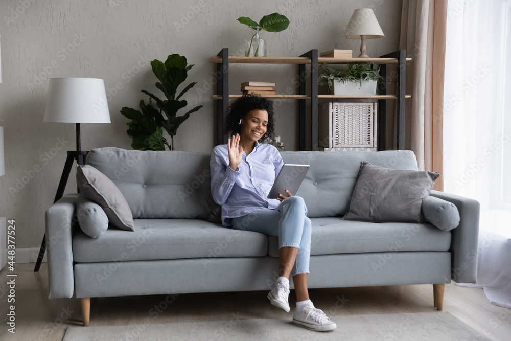 Smiling young African American woman sit on sofa at home wave greet talking speaking on video call on pad. Happy biracial female have webcam digital virtual event on tablet. Communication concept. Smiling young African American woman sit on sofa at home wave greet talking speaking on video call on pad. Happy biracial female have webcam digital virtual event on tablet. Communication concept.