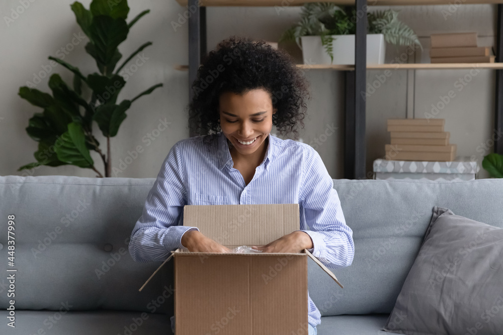 Excited young African American woman open unpack box shopping online from home. Smiling biracial female buyer client unbox package with internet order buy on web. Good delivery service concept. Excited young African American woman open unpack box shopping online from home. Smiling biracial female buyer client unbox package with internet order buy on web. Good delivery service concept.