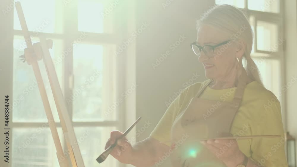 Senior Artist, Art Workshop, Sun Rays, Painting Woman. Happy senior woman artist paints a picture on canvas while standing in a sunny workshop