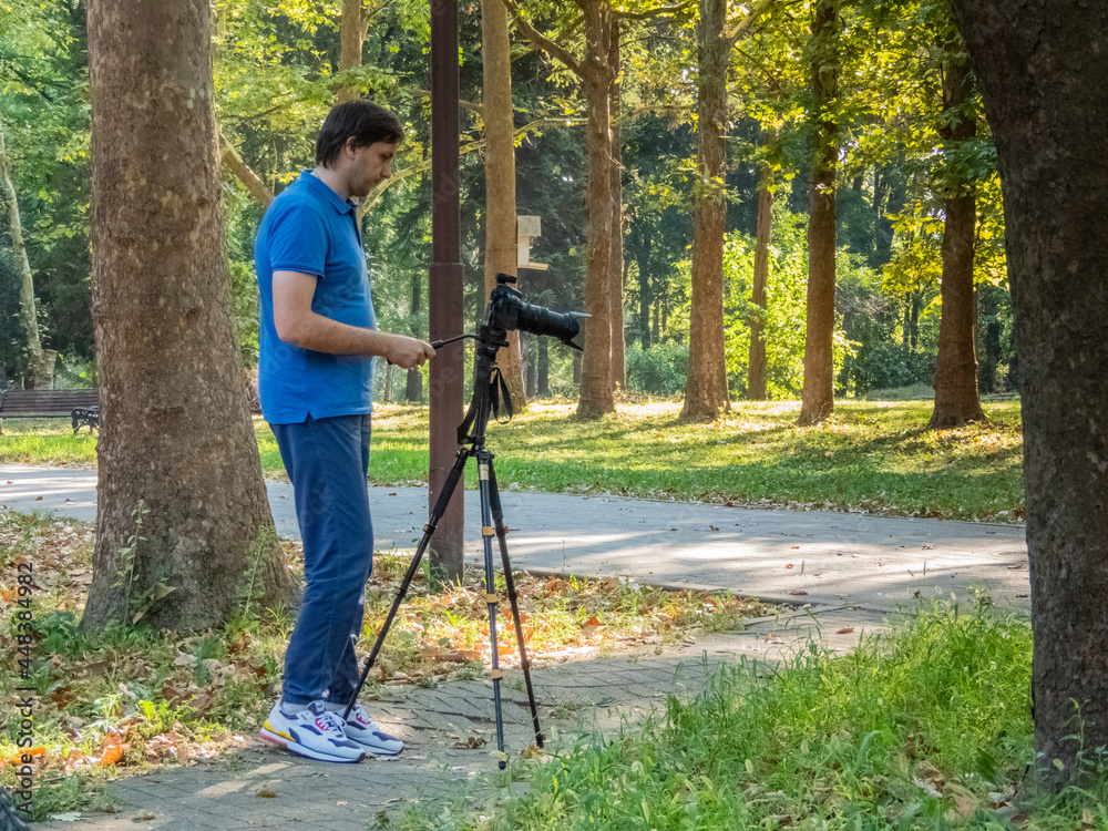 a young white male photographer with a blue t-shirt and a camera with a large lens mounted on a tripod in a park on a sunny summer day