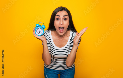Excited shouting young cute woman with a blue clock in hands isolated on yellow background © My Ocean studio