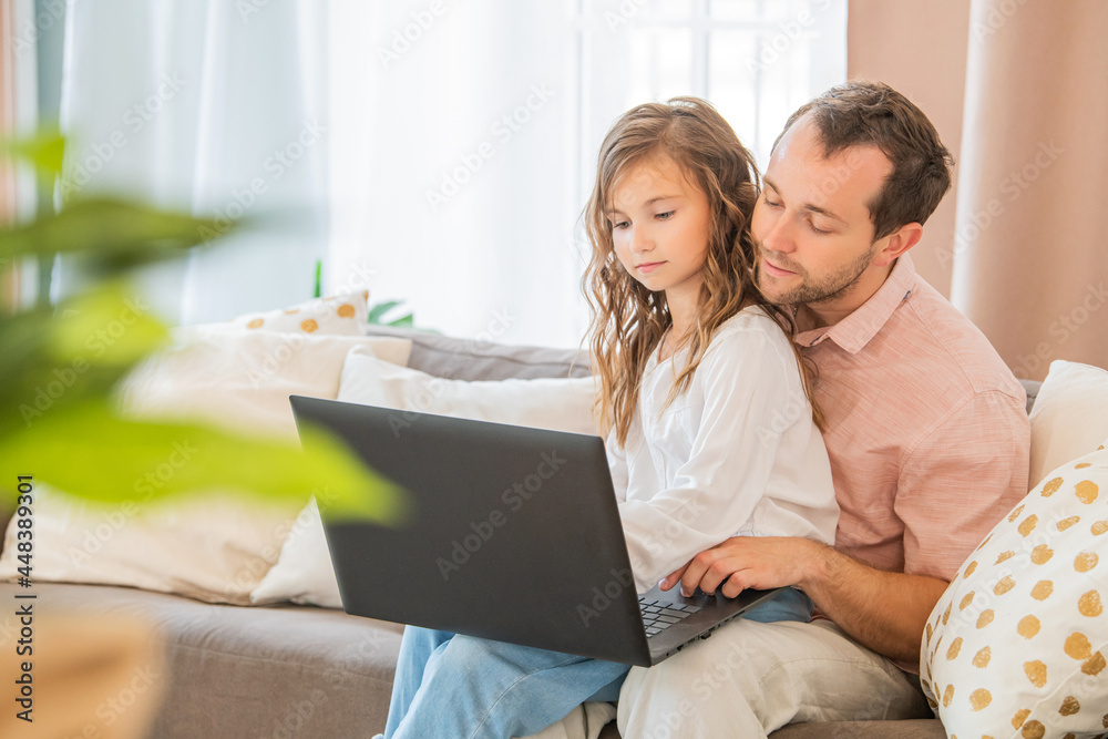 Happy family, daughter sitting on her father's lap on couch at home looking at laptop monitor together. Happy family, daughter sitting on her father's lap on couch at home looking at laptop monitor together.