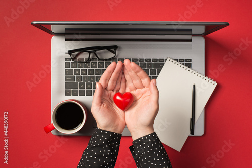 Overhead photo of grey laptop notepad pen glasses cup of coffee and hands holding a red heart isolated on the red backdrop photo