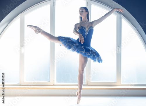 Focused young ballerina dressed in blue tutu costume makes pirouette at ballet studio in front of big round shaped window background in natural ambient daylight atmosphere