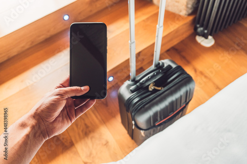 Man taking a smartphone and in the background his travel suitcase, just before leaving for the trip. It is located in a room of a hotel with wooden floor.