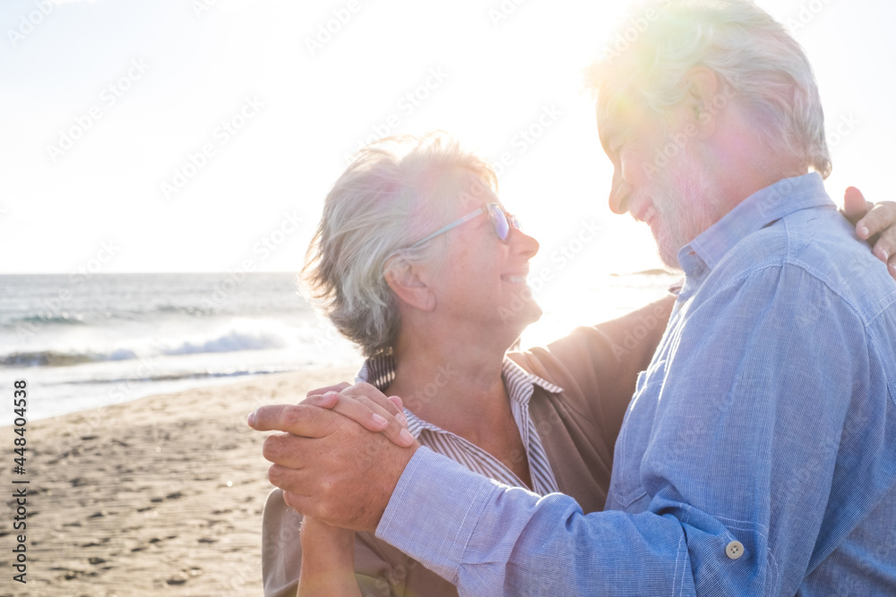 Couple of two old and mature happy seniors enjoying summer dancing together at the beach on the sand with the sunset at the background. Retired and leisure lifestyle.