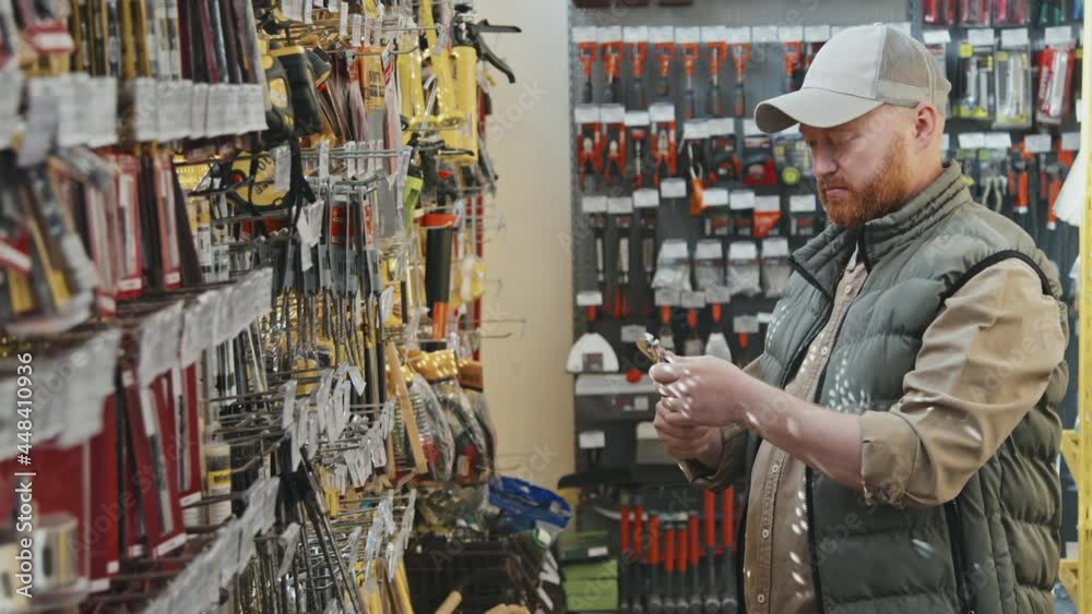Slowmo PAN of bearded man in puffy vest and cap looking at wrench at hardware store, then turning towards camera Slowmo PAN of bearded man in puffy vest and cap looking at wrench at hardware store, then turning towards camera