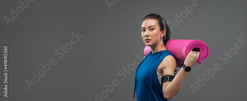 Portrait beautiful Asian woman sweating in sportswear blue color and holding yoga mat looking at camera after cardio exercise and standing on banner grey background.