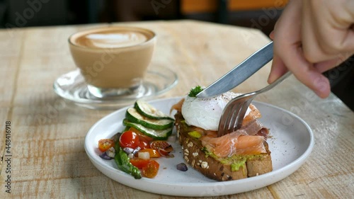 Healthy breakfast in cafe - benedict eggs with salmon and avocado on whole grain toast with grilled vegetables on plate. Man hand cutting fresh bread with poached egg in cafe. Concept of fitness menu photo