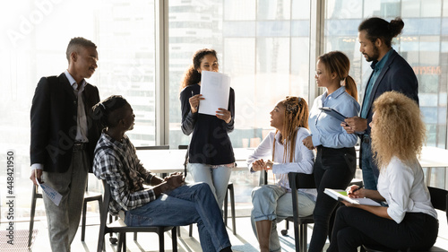 Happy shy young african american employee holding paper documents in hands, presenting marketing research results to interested multiracial diverse colleagues at brainstorming meeting in office.