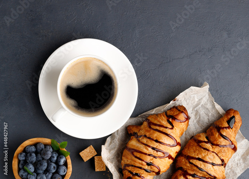 Breakfast - coffee, sweet croissant with chocolate and blueberries. Black stone background. Top view.