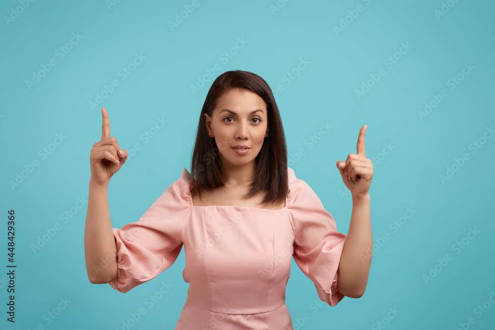 Caucasian woman with dark hair in pink dress has confident facial expression, indicates with both index fingers up, demonstrates copy space for your advertising content, isolated on blue background. Caucasian woman with dark hair in pink dress has confident facial expression, indicates with both index fingers up, demonstrates copy space for your advertising content, isolated on blue background.