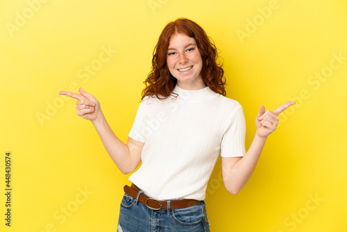 Teenager reddish woman isolated on yellow background pointing finger to the laterals and happy