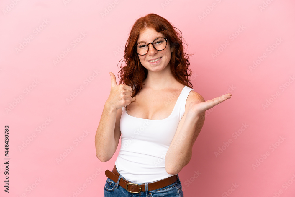 Teenager reddish woman isolated on pink background holding copyspace imaginary on the palm to insert an ad and with thumbs up Teenager reddish woman isolated on pink background holding copyspace imaginary on the palm to insert an ad and with thumbs up