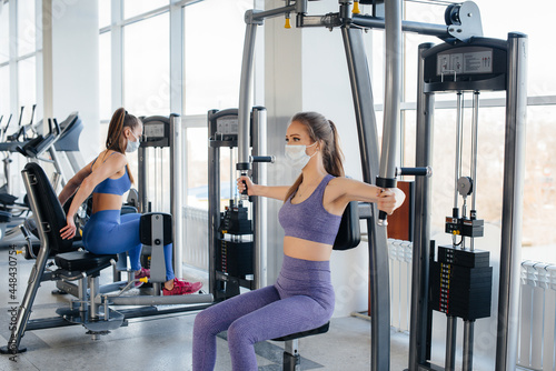 Two young beautiful girls exercise in the gym wearing masks during the pandemic. Social distancing in public places.