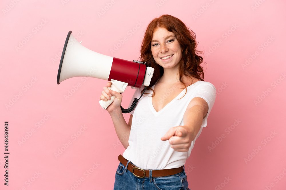Teenager reddish woman isolated on pink background holding a megaphone and smiling while pointing to the front Teenager reddish woman isolated on pink background holding a megaphone and smiling while pointing to the front