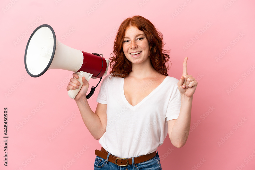 Teenager reddish woman isolated on pink background holding a megaphone and pointing up a great idea