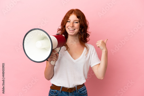 Teenager reddish woman isolated on pink background shouting through a megaphone and pointing side