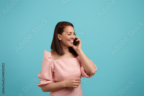 Free time. Joyful broadly smiling european woman with dark hair in pink dress talks on phone, enjoys conversation, looks away, has nice talk, keeps smartphone near ear, isolated on blue background.