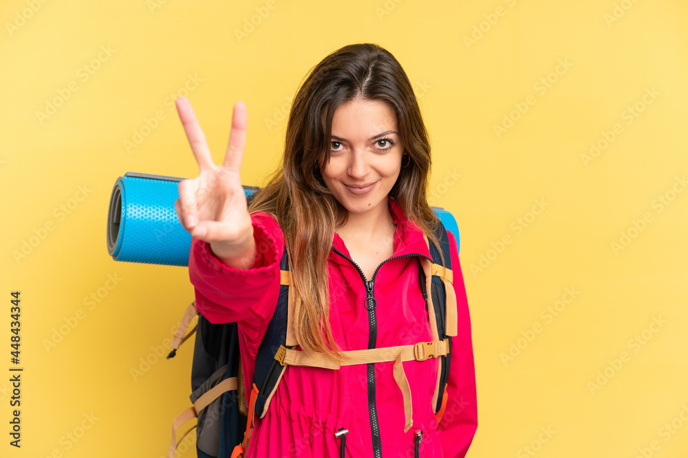 Young mountaineer man with a big backpack isolated on yellow background smiling and showing victory sign Young mountaineer man with a big backpack isolated on yellow background smiling and showing victory sign