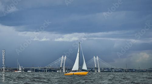 The yellow sailboat on the horizon in sea at sunset, cable bridge on background, the storm sky of different colors, big waves, sail regatta, cloudy weather