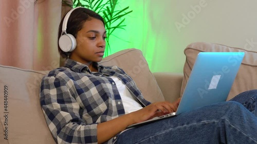 A young beautiful African-American woman wearing headphones lying at home on the couch won a big prize online. The concept of happiness, joy and luck.