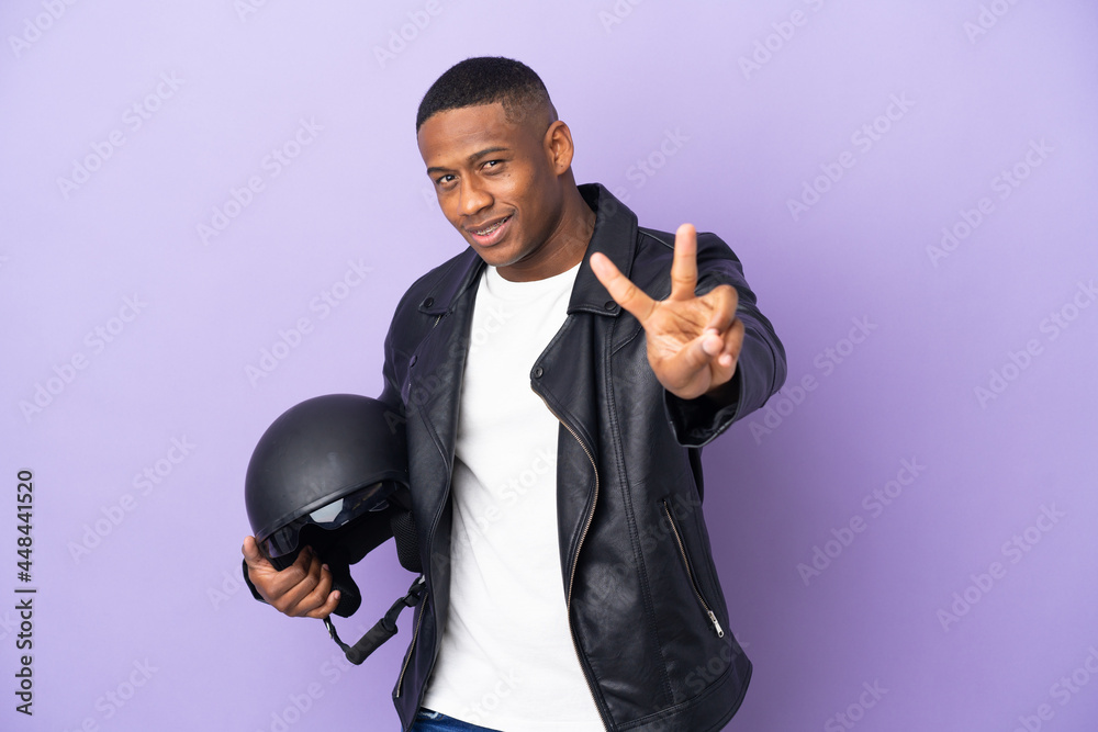 Latin man with a motorcycle helmet isolated on purple background smiling and showing victory sign Latin man with a motorcycle helmet isolated on purple background smiling and showing victory sign
