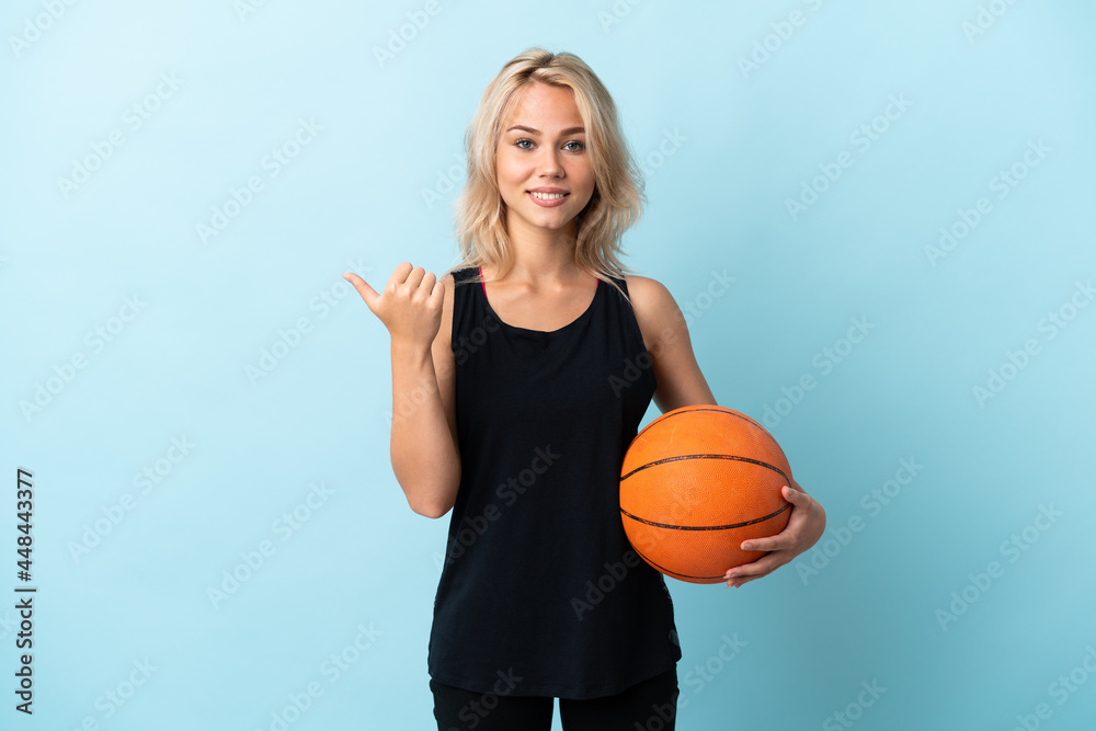 Young Russian woman playing basketball isolated on blue background pointing to the side to present a product