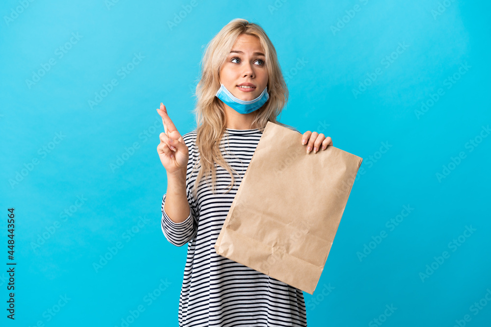 Young Russian woman holding a grocery shopping bag isolated on blue background with fingers crossing and wishing the best Young Russian woman holding a grocery shopping bag isolated on blue background with fingers crossing and wishing the best