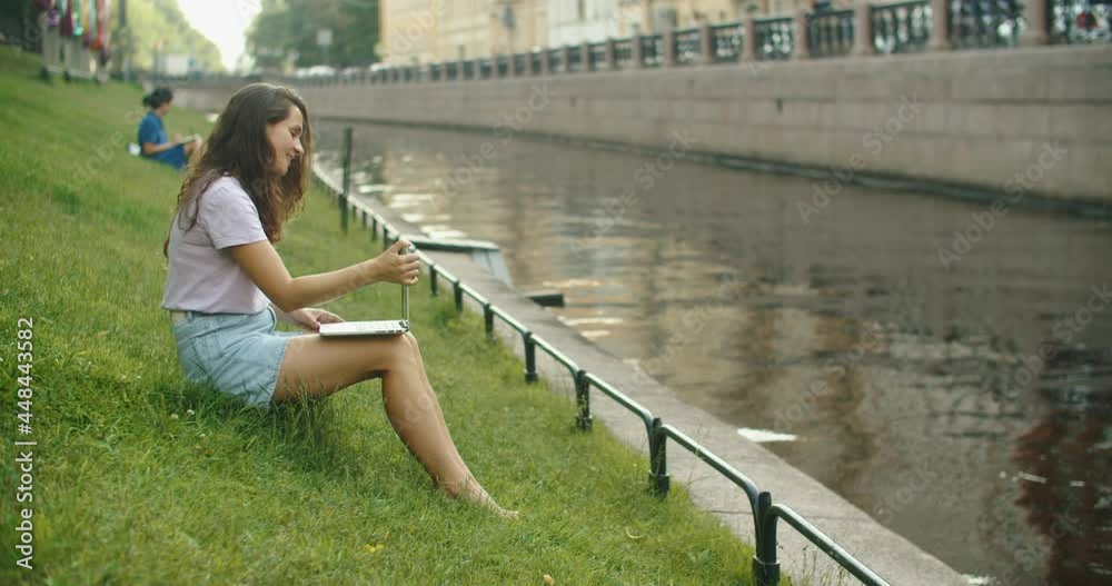 Happy brunette woman working on laptop outdoors sitting on the grass in the park. Nice place to work. Freelance. High quality 4k footage Happy brunette woman working on laptop outdoors sitting on the grass in the park. Nice place to work. Freelance. High quality 4k footage