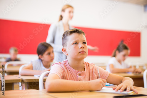 Boy and his classmates sitting at desks in classroom. Their teacher standing in background.