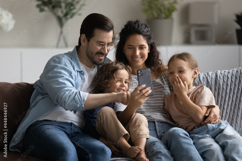 Happy family couple and little kids using mobile phone, sitting together on couch at home, taking selfie, making video call. Parents and children looking at smartphone screen, laughing Happy family couple and little kids using mobile phone, sitting together on couch at home, taking selfie, making video call. Parents and children looking at smartphone screen, laughing