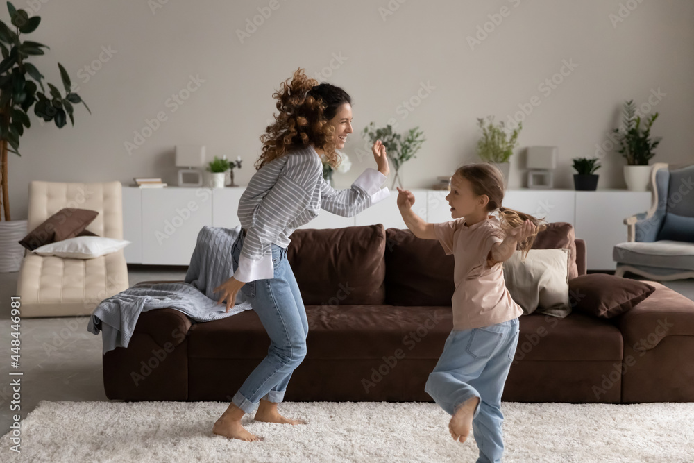 Happy excited young mom and little girl dancing to music in living room, enjoying home fitness exercises, playing active games, moving bodies, jumping, listening to radio, having fun Happy excited young mom and little girl dancing to music in living room, enjoying home fitness exercises, playing active games, moving bodies, jumping, listening to radio, having fun