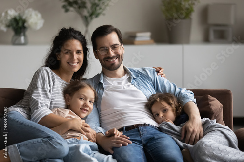 Happy family couple and two little kids resting on comfortable sofa at home. Portrait of young parents, son and daughter relaxing on couch, hugging, looking at camera, smiling. Homeowners, parenthood
