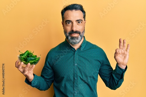 Middle age man with beard and grey hair holding green apples doing ok sign with fingers, smiling friendly gesturing excellent symbol