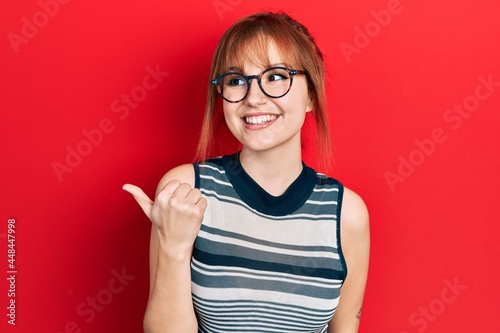 Fototapeta Naklejka Na Ścianę i Meble -  Redhead young woman wearing casual clothes and glasses smiling with happy face looking and pointing to the side with thumb up.