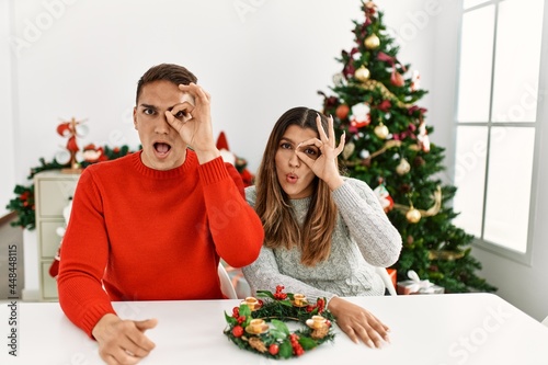 Young hispanic couple sitting at the table on christmas doing ok gesture shocked with surprised face, eye looking through fingers. unbelieving expression.