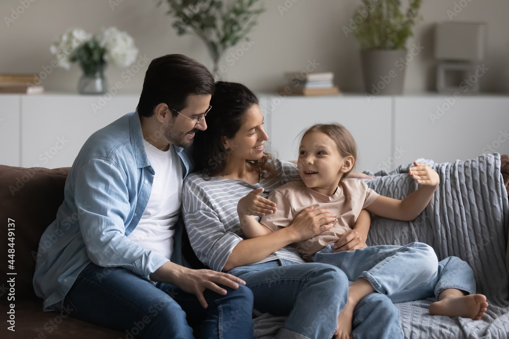 Happy excited daughter girl talking to engaged parents, telling funny story. Young family couple and child sitting together on couch, hugging, enjoying communication, leisure time together Happy excited daughter girl talking to engaged parents, telling funny story. Young family couple and child sitting together on couch, hugging, enjoying communication, leisure time together