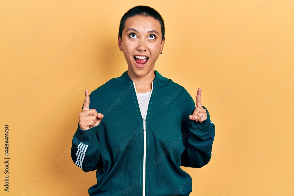 Beautiful hispanic woman with short hair wearing sporty jacket amazed and surprised looking up and pointing with fingers and raised arms. Beautiful hispanic woman with short hair wearing sporty jacket amazed and surprised looking up and pointing with fingers and raised arms.