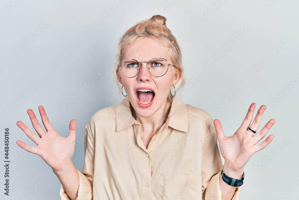 Beautiful caucasian woman with blond hair wearing casual look and glasses crazy and mad shouting and yelling with aggressive expression and arms raised. frustration concept. Beautiful caucasian woman with blond hair wearing casual look and glasses crazy and mad shouting and yelling with aggressive expression and arms raised. frustration concept.