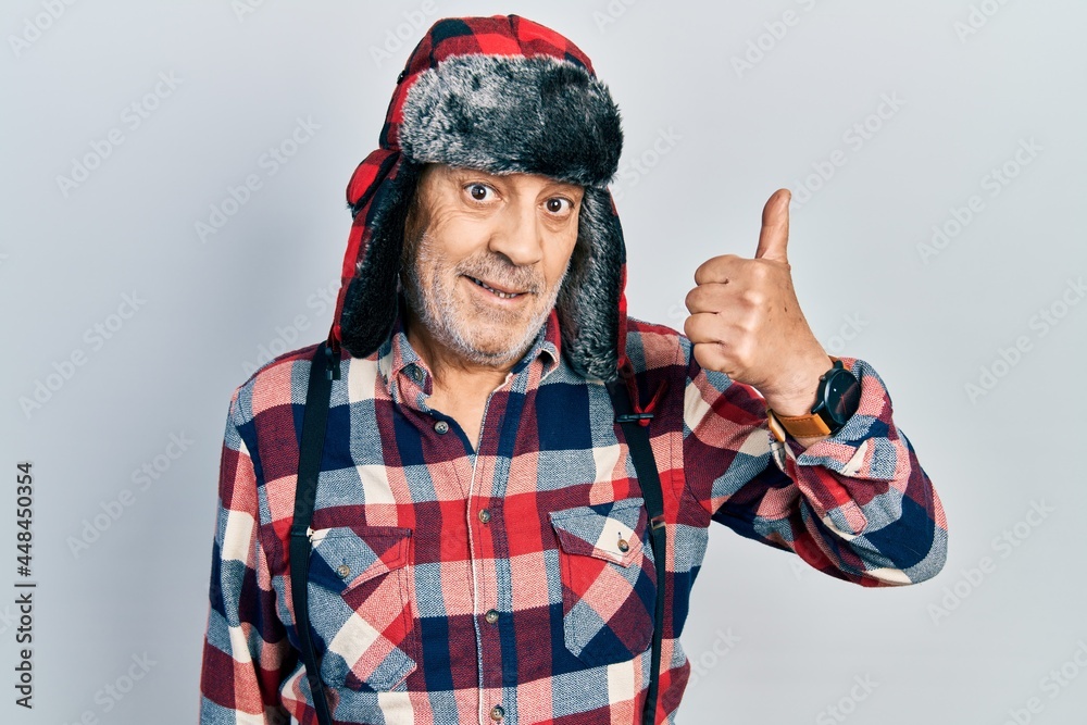Naklejka premium Handsome mature man wearing winter hat with ear flaps smiling happy and positive, thumb up doing excellent and approval sign