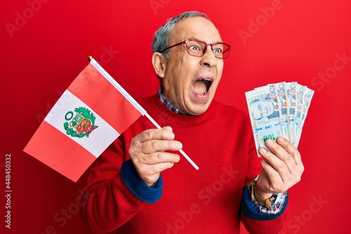 Handsome senior man with grey hair holding peru flag and peruvian sol banknotes angry and mad screaming frustrated and furious, shouting with anger. rage and aggressive concept.