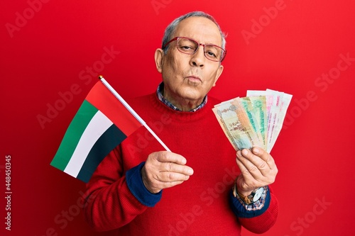Handsome senior man with grey hair holding united arab emirates dirham banknotes looking at the camera blowing a kiss being lovely and sexy. love expression.