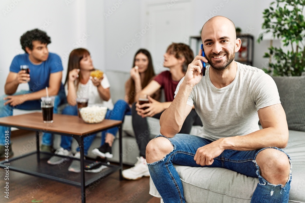 Group of young friends having party sitting on the sofa at home. Man smiling happy talking on the smartphone at home. Group of young friends having party sitting on the sofa at home. Man smiling happy talking on the smartphone at home.