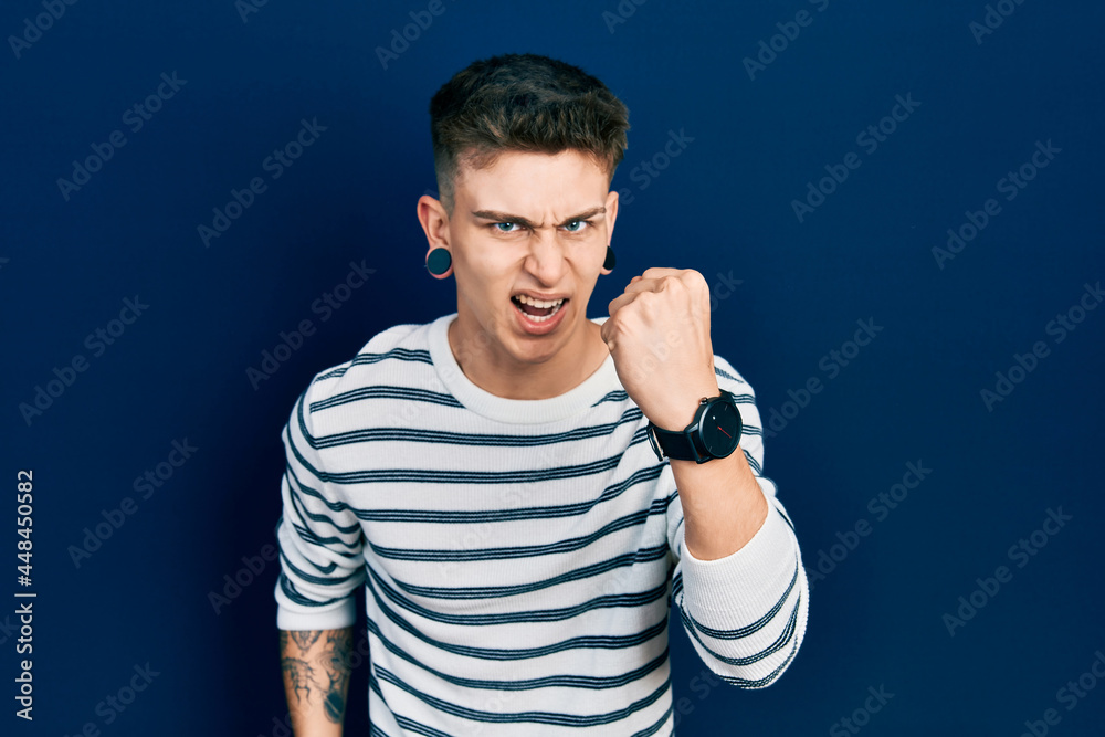 Young caucasian boy with ears dilation wearing casual striped shirt angry and mad raising fist frustrated and furious while shouting with anger. rage and aggressive concept. Young caucasian boy with ears dilation wearing casual striped shirt angry and mad raising fist frustrated and furious while shouting with anger. rage and aggressive concept.