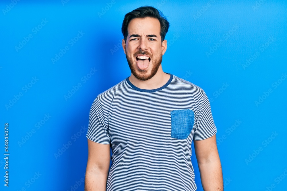 Handsome caucasian man with beard wearing casual striped t shirt sticking tongue out happy with funny expression. emotion concept. Handsome caucasian man with beard wearing casual striped t shirt sticking tongue out happy with funny expression. emotion concept.