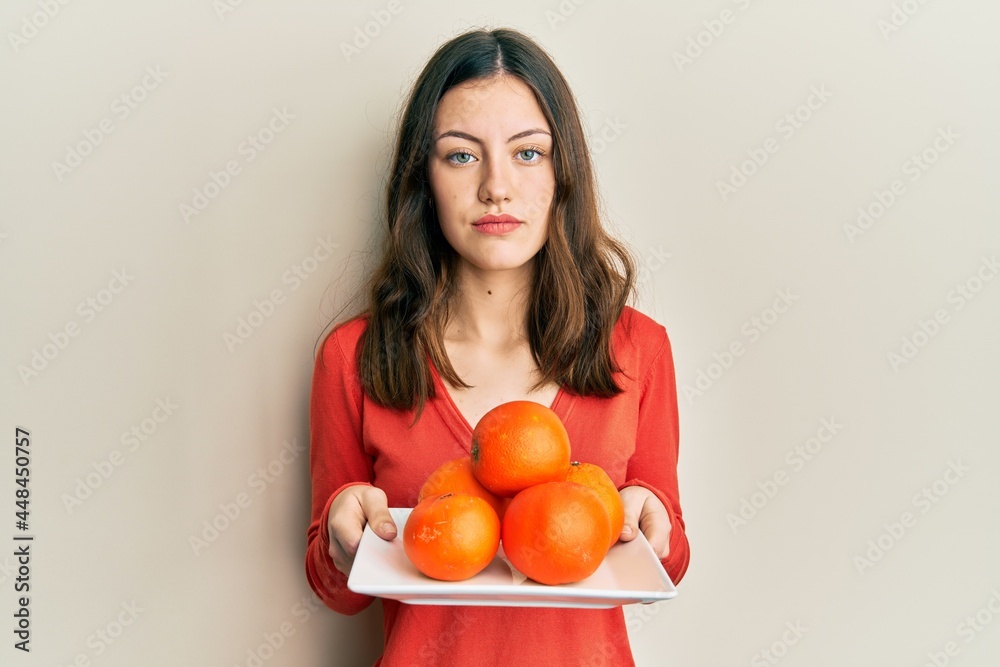 Young brunette woman holding plate with fresh oranges relaxed with serious expression on face. simple and natural looking at the camera. Young brunette woman holding plate with fresh oranges relaxed with serious expression on face. simple and natural looking at the camera.