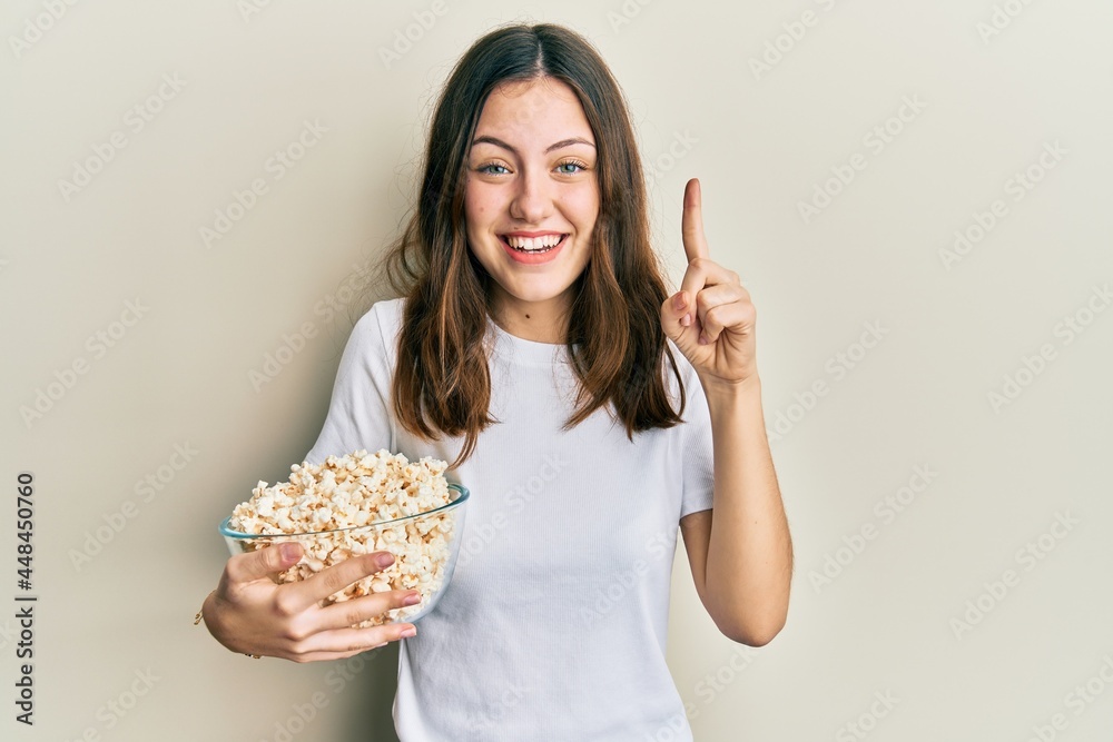 Young brunette woman eating popcorn smiling with an idea or question pointing finger with happy face, number one Young brunette woman eating popcorn smiling with an idea or question pointing finger with happy face, number one